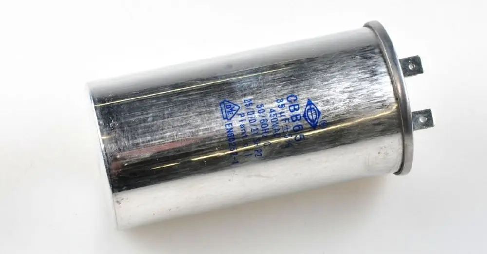 Kondensator für Kompressor, metallische Oberfläche mit Anschlussstiften, beschriftet mit technischen Angaben. Produkt von Remko für elektrische Anwendungen.