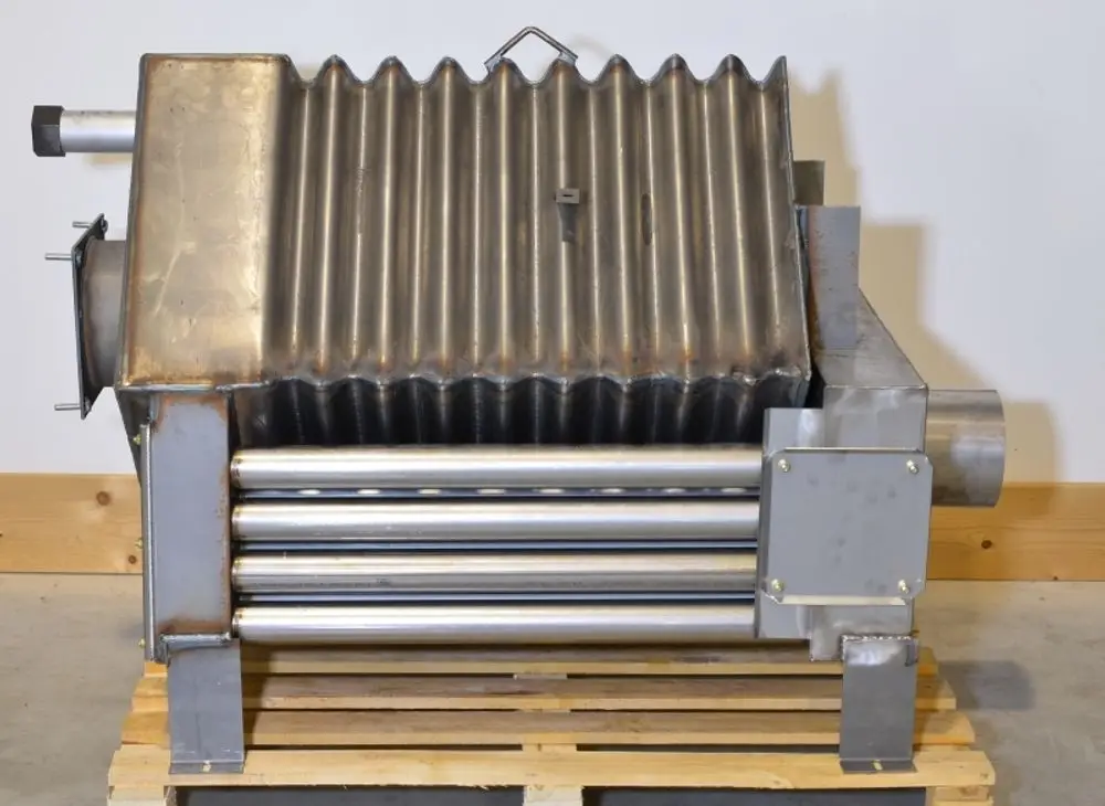 Brennkammer aus Edelstahl mit gerippter Oberfläche, montiert auf einer Holzpalette, zu sehen im Innenraum. Teile aus Metall, unter anderem Rohre zur Wärmeleitung. Hersteller: Remko.