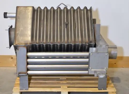 Brennkammer aus Edelstahl mit gerippter Oberfläche, montiert auf einer Holzpalette, zu sehen im Innenraum. Teile aus Metall, unter anderem Rohre zur Wärmeleitung. Hersteller: Remko.
