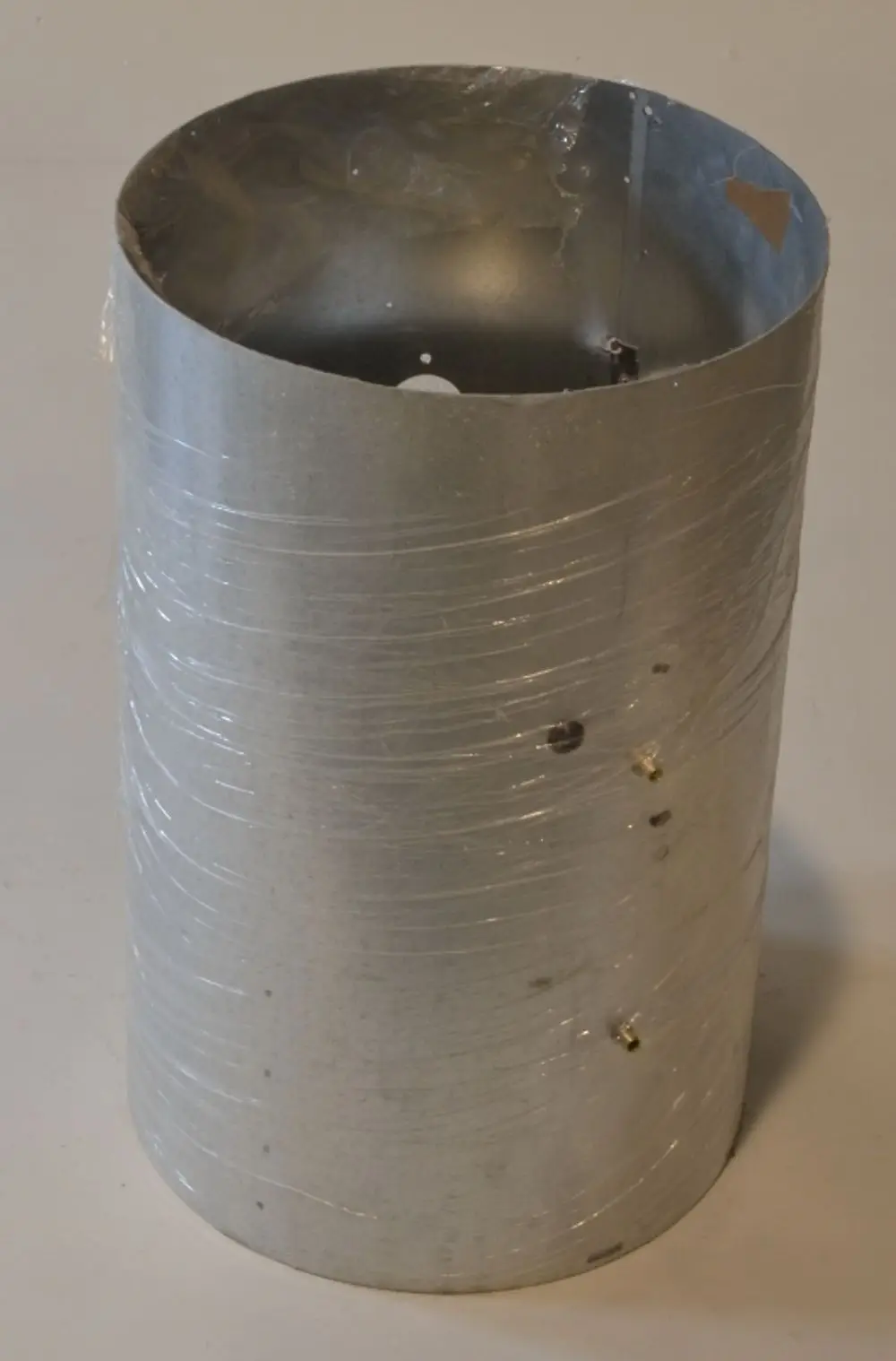 Einfache silberne Metallbrennkammer der Marke Remko, zylindrisch mit einer transparenten Folie umwickelt, an der Oberseite offen und mit mehreren Löchern an der Seitenfläche.