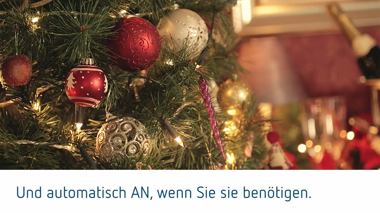 Weihnachtlich dekorierter Baum mit verschiedenen farbigen Ornamenten und Lichtern. Im Hintergrund Gläser und Sektflasche. Die Szene vermittelt eine festliche Atmosphäre.