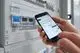 Person hält ein Smartphone, das eine App zeigt, während im Hintergrund ein Schaltkasten mit Theben-Geräten sichtbar ist. Die Geräte haben digitale Anzeigen und Tasten.