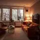 Wohnzimmer mit einer orangen Couch, einem kleinen Tisch und einem Fernseher. Große Fenster mit Blick auf schneebedeckte Bäume. Pflanzen und eine Lampe ergänzen die Einrichtung.