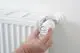 Hand betätigt einen weißen Thermostatkopf von Danfoss, der an einem Heizkörper montiert ist. Der Heizkörper ist horizontal und läuft parallel zur Wand.
