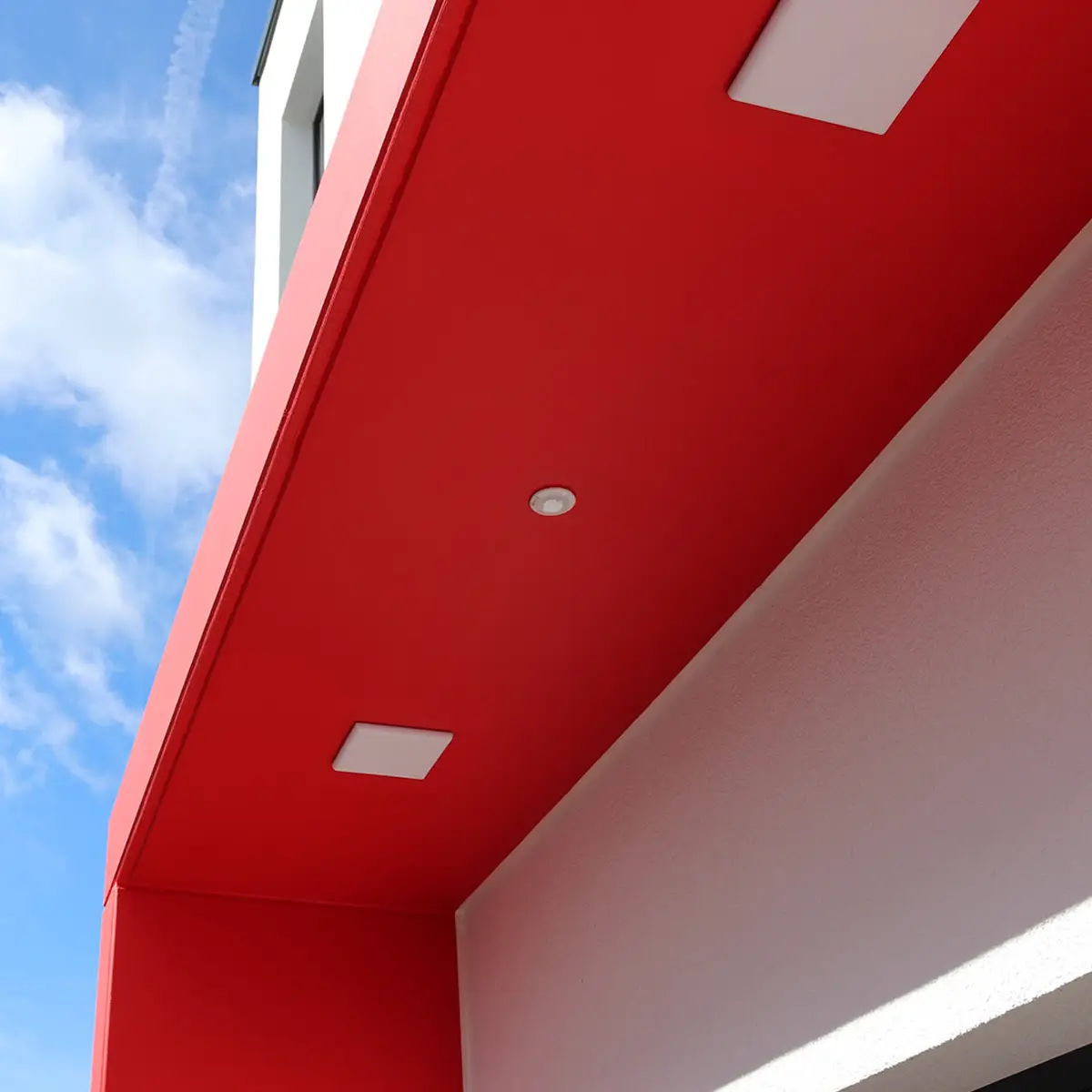 Architekturdetail: Rotes Vordach mit weißen Lampen und Fensterausschnitt vor blauem Himmel.