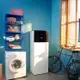 Weißes Innengerät von Daikin Altherma 4, auf einer grauen Bodenfliesenfläche, neben einer Waschmaschine und einem Kühlschrank, in einem Raum mit blauer Wand und Fenster.