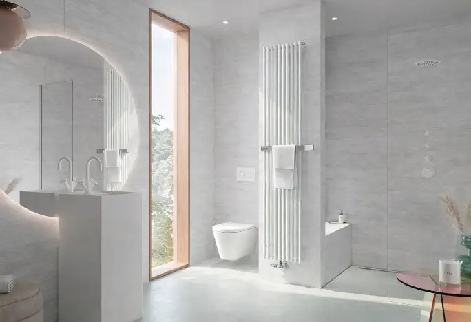 Helles Badezimmer mit Kermi Pio plus Heizkörper, weiß, Handtuchbügel aus Edelstahl, neben Fenster und Toilette. Runder Spiegel über modernem Waschbecken.