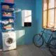 Wandgerät von Daikin in Weiß an blauer Wand, daneben ein Waschmaschine und ein Fahrrad. Regalsystem mit Handtüchern und einer Lampe auf einem Fensterbrett.
